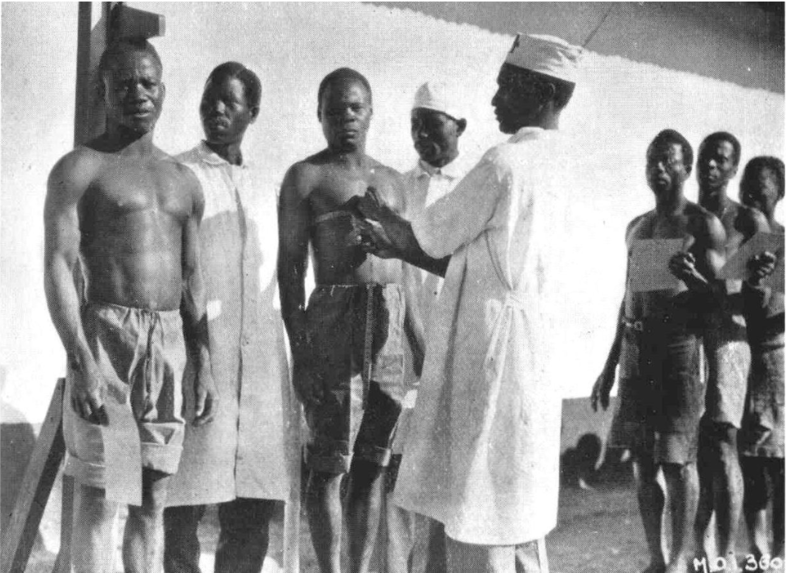 Historical photograph of a medical examination of Congolese workers at a UMHK camp: a medical officer in white coat examines a line of men stripped to the waist.